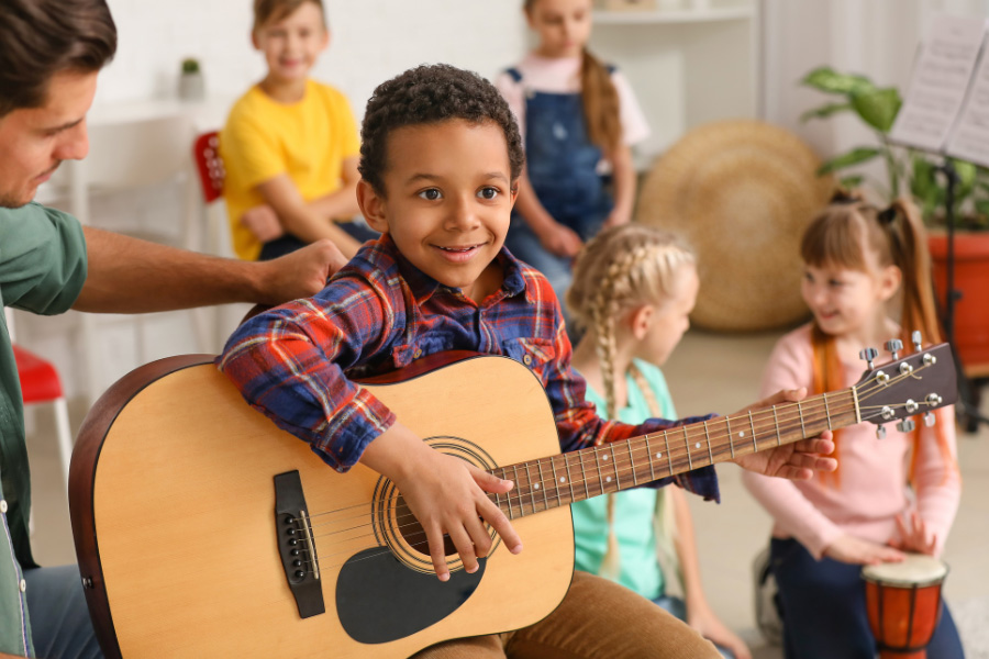 young boy learning to play guitar with other children playing percussion instruments